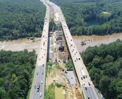 Aerial view of a divided highway with ongoing construction in the center, surrounded by dense green forest and a river running parallel to the road.
