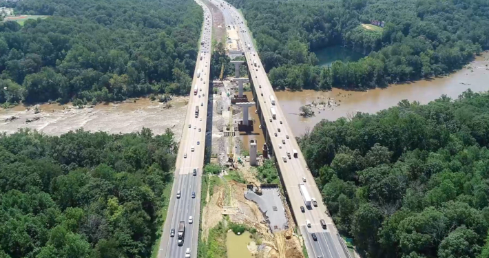 Aerial view of a divided highway with ongoing construction in the center, surrounded by dense green forest and a river running parallel to the road.
