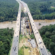 Aerial view of a divided highway with ongoing construction in the center, surrounded by dense green forest and a river running parallel to the road.