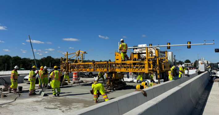 Construction workers in neon vests operate machinery on a highway under a clear blue sky. They are finishing a new concrete pour on the road using large equipment.