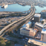 Aerial view of a city with highways, overpasses, office buildings, and a waterfront with ships docked in the background.