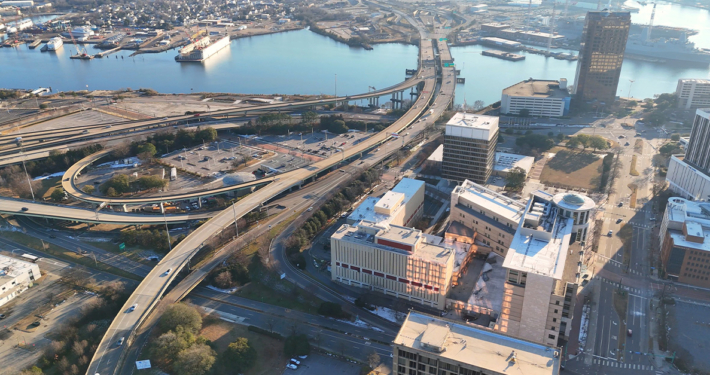 Aerial view of a city with highways, overpasses, office buildings, and a waterfront with ships docked in the background.