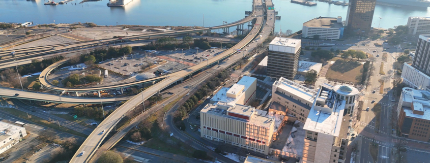 Aerial view of a city with highways, overpasses, office buildings, and a waterfront with ships docked in the background.