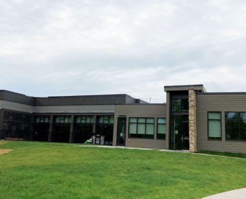 Modern gray community building with large windows, set on a green lawn under a cloudy sky. A stone accent marks the entrance.