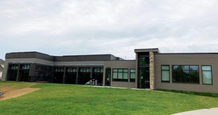 Modern gray community building with large windows, set on a green lawn under a cloudy sky. A stone accent marks the entrance.