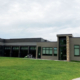 Modern gray community building with large windows, set on a green lawn under a cloudy sky. A stone accent marks the entrance.