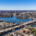 Aerial view of a city with a wide river, two bridges, and surrounding residential areas. The clear blue sky conveys a calm, sunny ambiance.