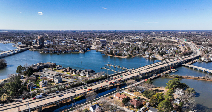 Aerial view of a city with a wide river, two bridges, and surrounding residential areas. The clear blue sky conveys a calm, sunny ambiance.