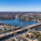 Aerial view of a city with a wide river, two bridges, and surrounding residential areas. The clear blue sky conveys a calm, sunny ambiance.