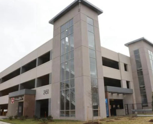 Large multi-story parking garage with tall glass-panel stair towers and open parking levels. Modern, urban design with a neutral tone under a cloudy sky.
