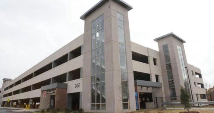 Large multi-story parking garage with tall glass-panel stair towers and open parking levels. Modern, urban design with a neutral tone under a cloudy sky.
