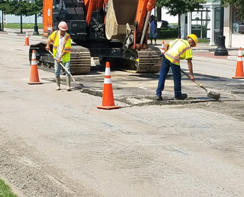 Construction workers actively working on a street in front of a building, using tools and machinery for repairs.