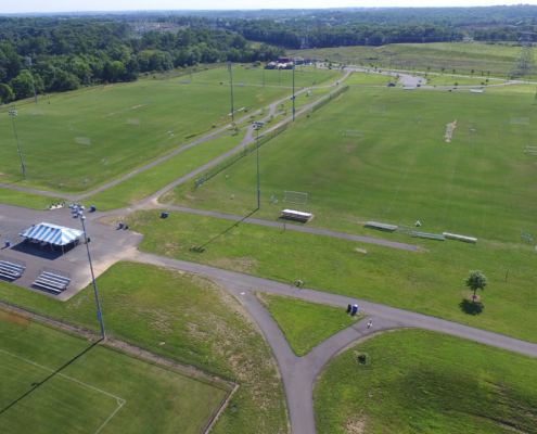 Aerial view of a vast, green sports complex with multiple soccer fields. Bleachers and a tent are visible; surrounded by trees under a clear sky.