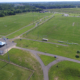 Aerial view of a vast, green sports complex with multiple soccer fields. Bleachers and a tent are visible; surrounded by trees under a clear sky.